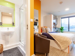 A modern bedroom with a large bed, a bedside table, a chair, a window, and a bathroom with a sink and mirror. - Student Only Hannah Court (Sheffield)