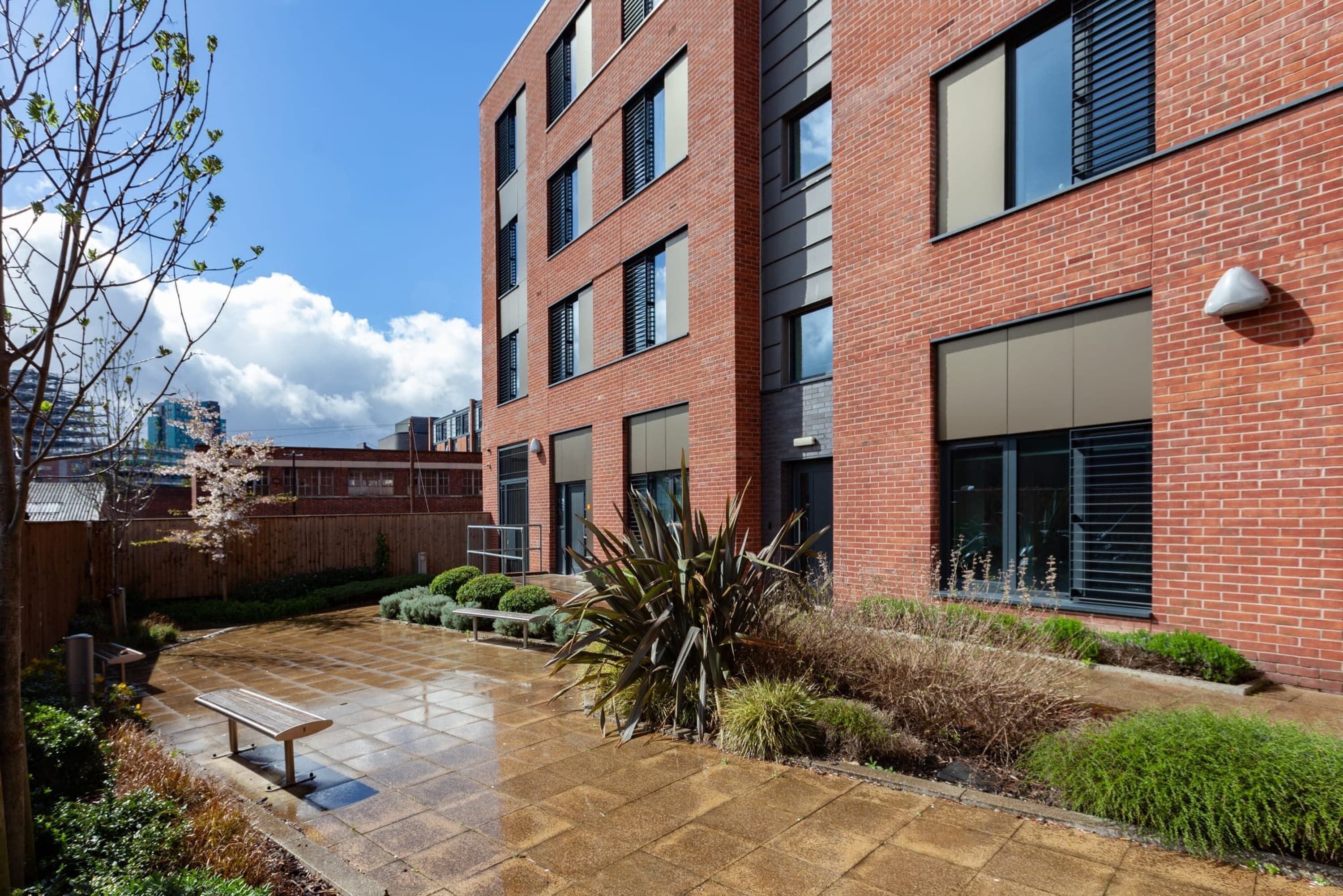 A multi-story brick building with a courtyard featuring a bench, potted plants, and a water feature.