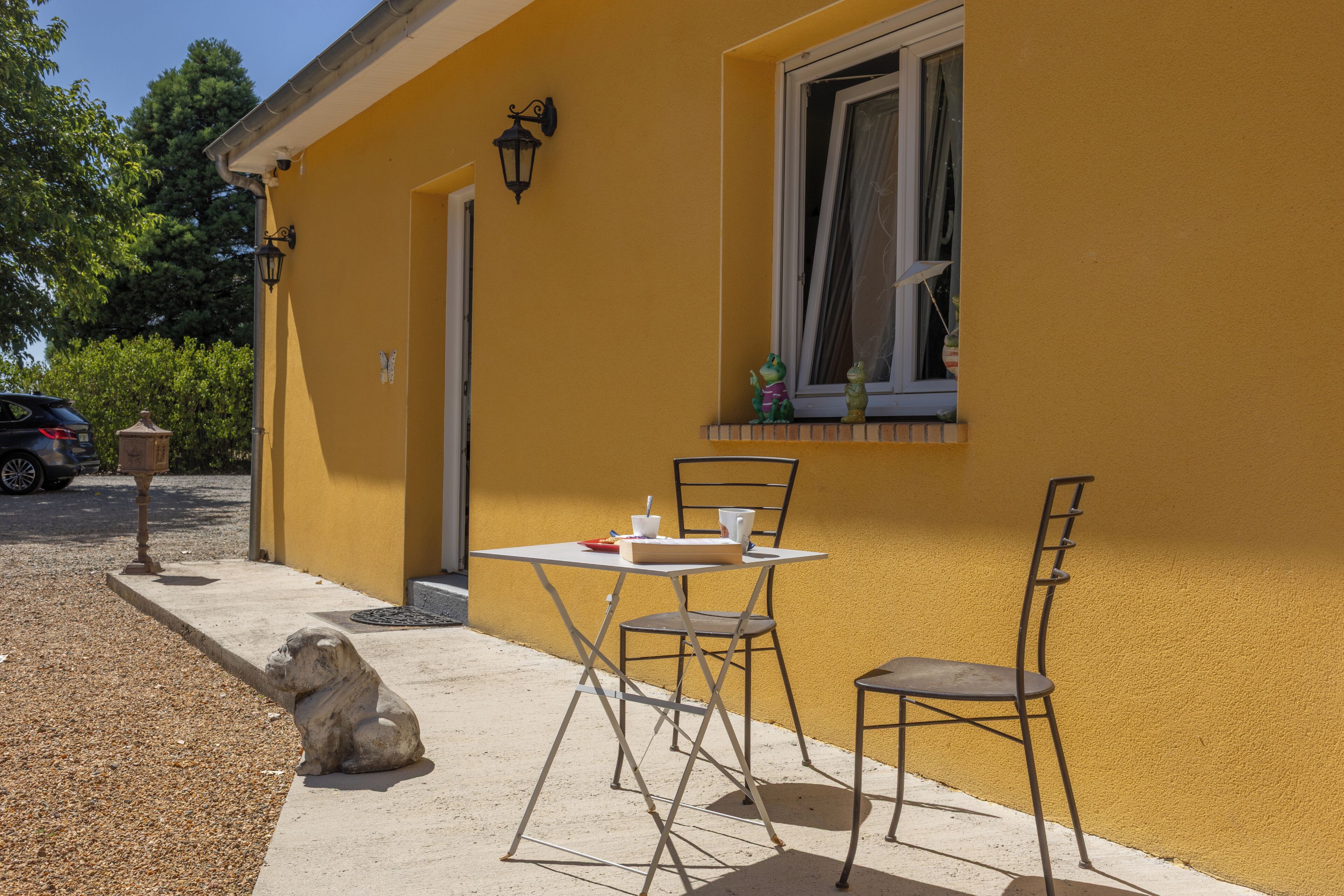 A yellow building with a window, a table with chairs, and a stone sculpture on the ground.