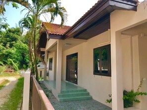 Una casa de dos pisos con un porche cubierto, una escalera y vistas del entorno verde.