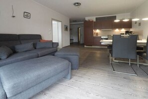 A modern living room with a grey sectional sofa, a dining area with a table and chairs, and a kitchen with wooden cabinets and a countertop. - Holiday apartment with 2 bedrooms (Dierhagen)