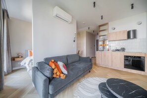 A modern living room with a grey sofa, a small table, and a kitchen area with wooden cabinets and a built-in oven. - Kierbedzia Boutique Studio, Mokotów, Warsaw (Warszawa)