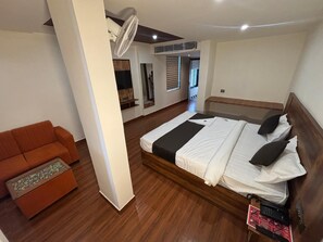 A hotel room with a bed, a sofa, a standing fan, and a wooden floor. - Hotel Darbaar Inn (Jaipur)
