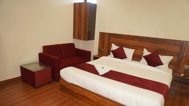 Habitación de hotel con cabecera de madera, sofá rojo, una mesa de centro de vidrio y cama con ropa blanca y detalles rojos.