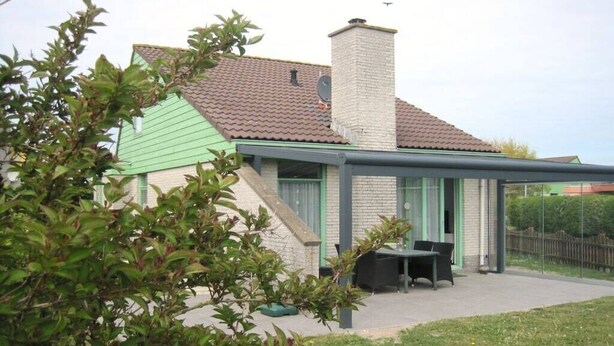 Uma casa com telhado verde, chaminé de tijolos e varanda coberta com mesa e cadeiras.
