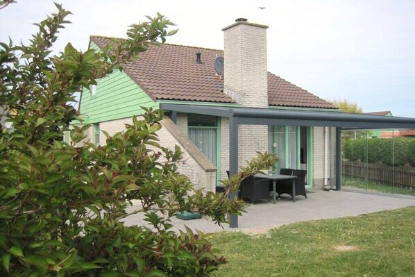 A house with a green roof, a brick chimney, and a covered patio area with a table and chairs. - Holiday home with 3 bedrooms (Julianadorp)