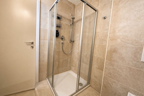 A glass-enclosed shower stall with a showerhead and control panel, beige tiled walls, and a door with a handle. - Vrbo Property (Kaprun)