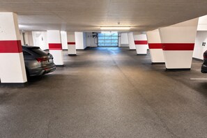 Uma garagem subterrânea com pilares de concreto, um carro cinza estacionado e céu visível pelas janelas.