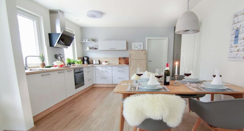 A modern kitchen with white cabinets, a wooden dining table set for four, and a large window providing natural light.