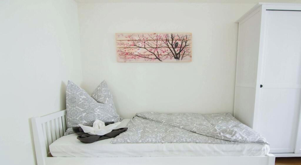 Une petite chambre blanche avec un lit simple, une tête de lit blanche, un oreiller à motifs gris et une couverture grise.