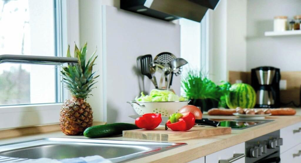Una cocina con una ventana, un colador, una piña, un pepino, pimientos rojos y una tabla de cortar.