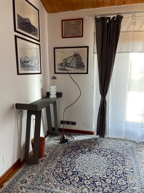 A room with a patterned rug, a console table with a vase, framed pictures on the wall, and a window with curtains. - Relais Infinito: Rooms and Views of the Marche Hills (Montefiore)
