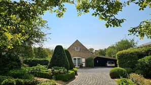 Une maison avec une allée pavée menant à un garage, entourée de haies bien entretenues et de diverses espèces d’arbustes.