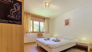 A bedroom with a bed, a chair, a nightstand, a window with curtains, and a poster on the wall. - Résidence Mila n°2 by Interhome (Crans-Montana)