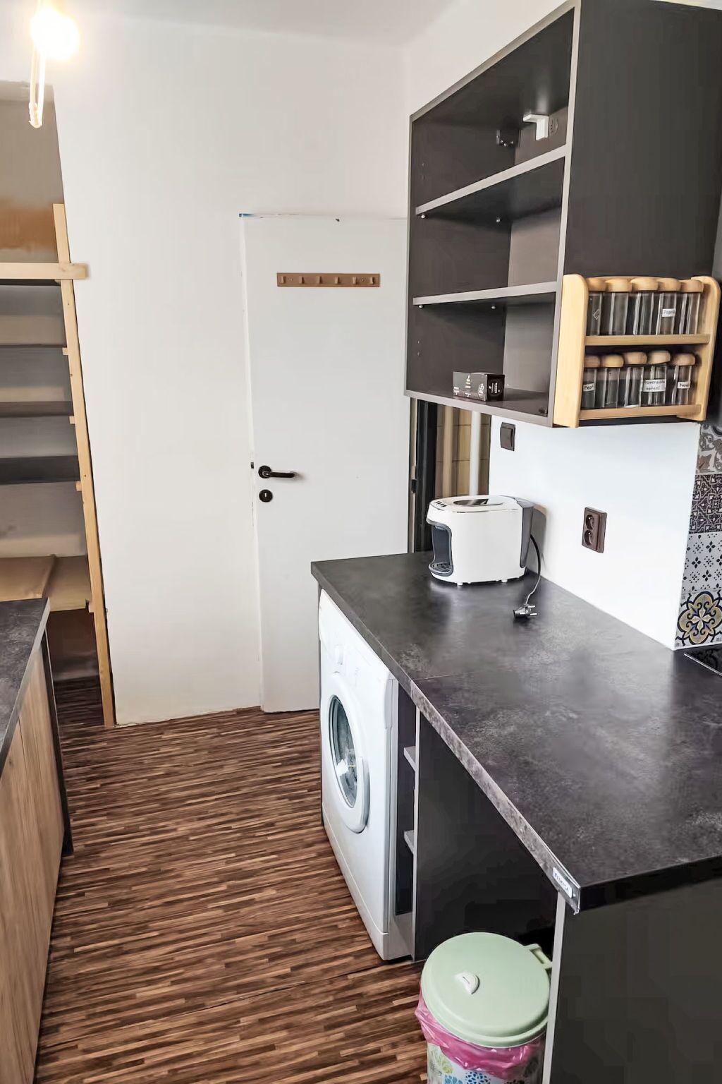 Een compacte keuken met een wasmachine, een koffiezetapparaat en een inbouwkast met potten.