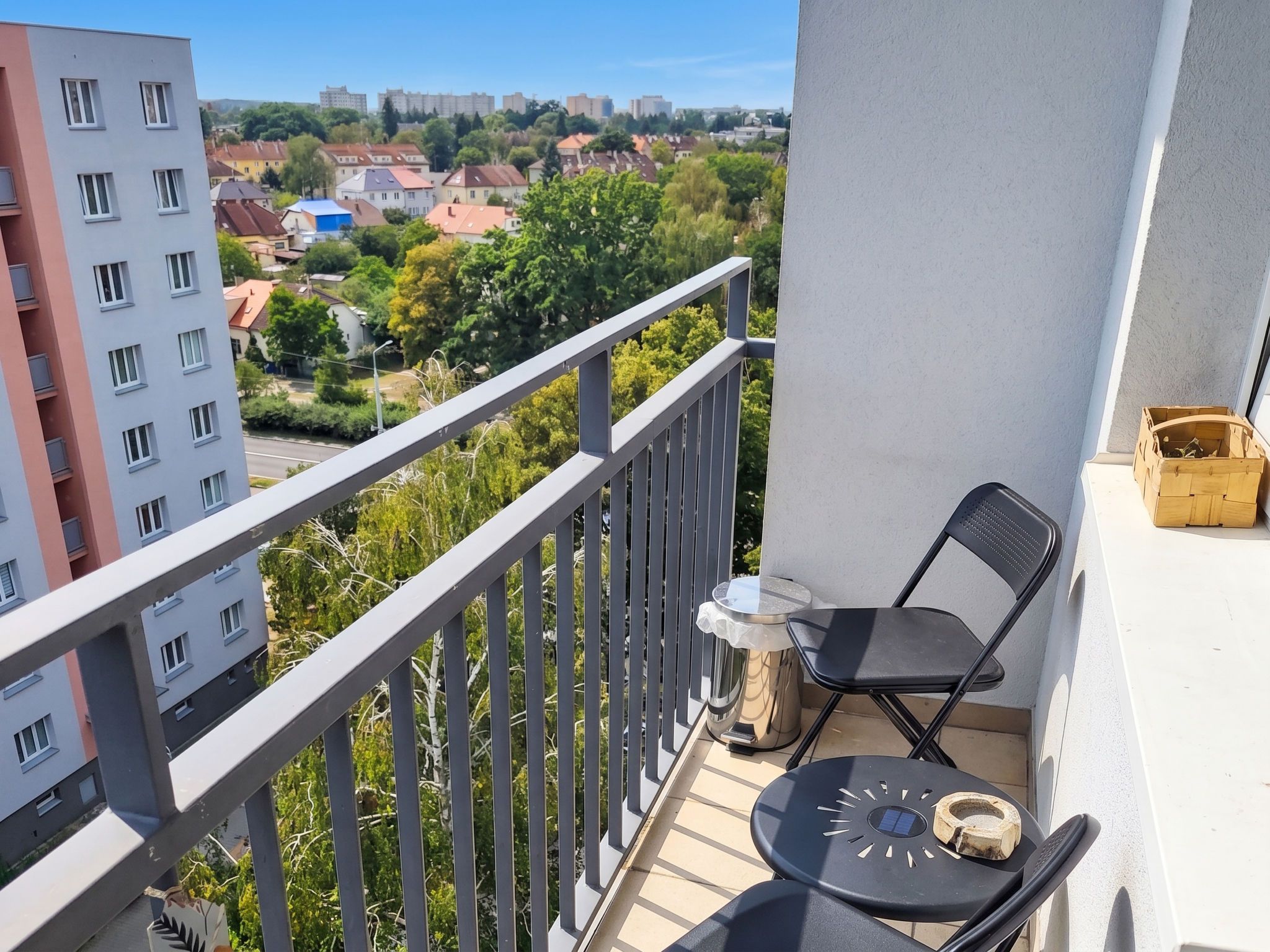 Un balcón con vistas a un paisaje urbano, una mesa con una silla y un pequeño contenedor de madera.