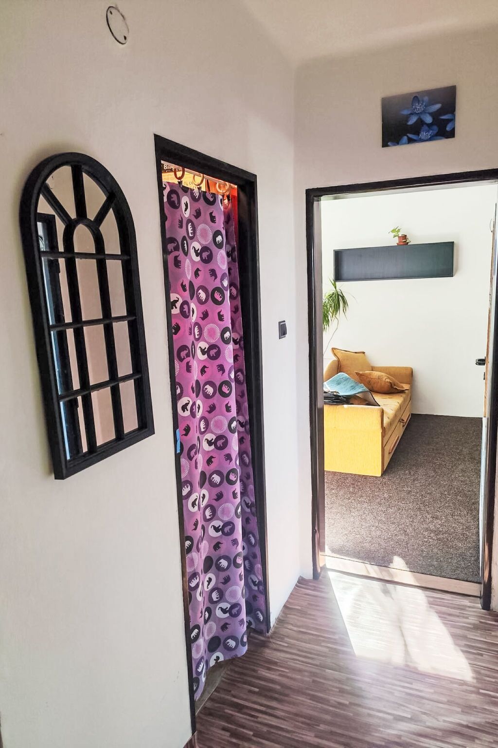 Un vano con un cortinero estampado, una ventana de marco negro y un cuarto con un sofá y una planta.