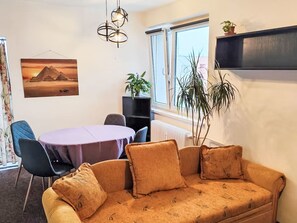 A cozy room with a sofa, a round table, and chairs. There's a window with curtains, a radiator, and a wall-mounted shelf with a plant.