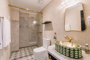 Et badeværelse med en glasvæg omkring bruseniche, et hvidt toilet, en grøn vask med guldfarmet armatur og et spejl med gylden ramme.