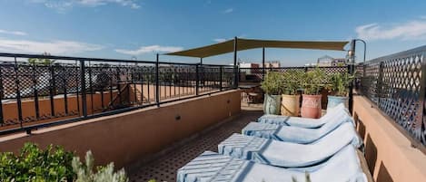 A rooftop terrace with lounge chairs, potted plants, and a table under a shaded area.
