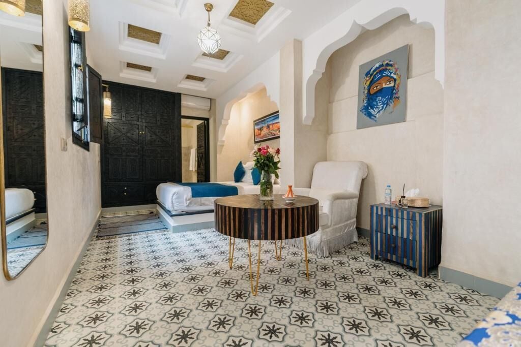 Habitación de hotel con piso estampado, una mesita redonda, un sillón blanco y un cuadro en la pared.