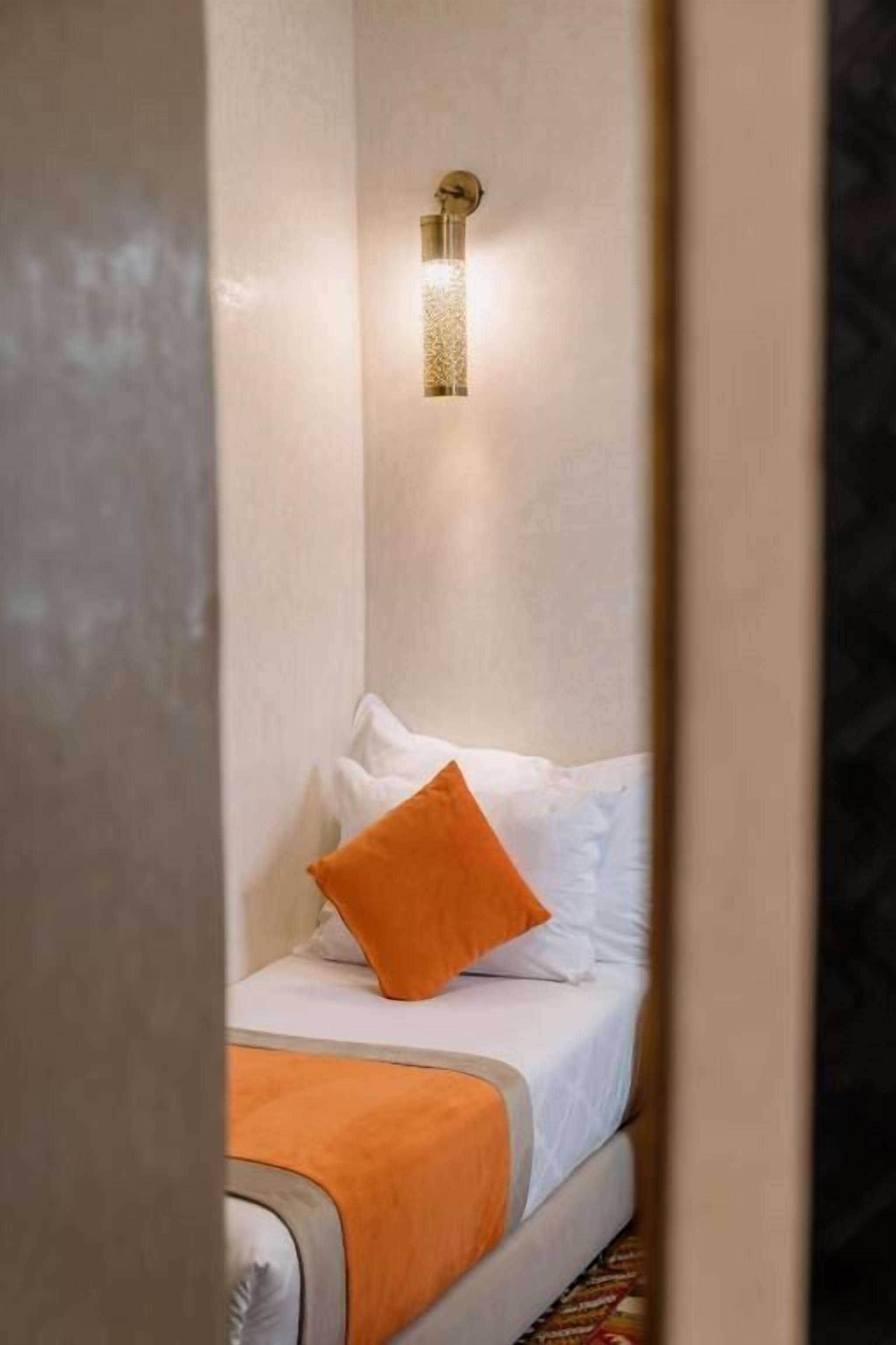 Habitación de hotel con cama, almohada anaranjada, lámpara de pared y pared con textura.