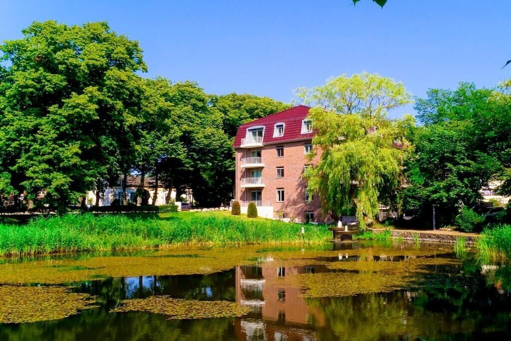 A multi-story building with a red roof, surrounded by lush greenery and a pond with lily pads.