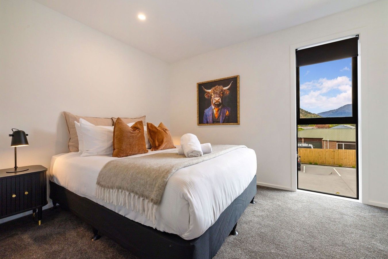 A bedroom with a large bed, a nightstand with a lamp, a painting of a bull, and a view of a parking lot through a sliding door.