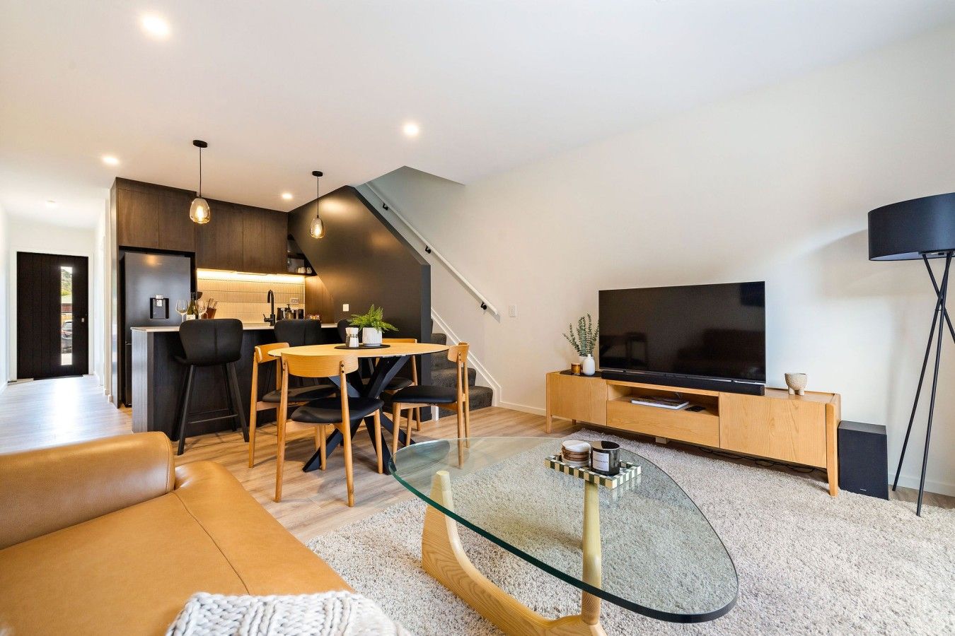 A modern living room with a glass coffee table, a sofa, a dining area with chairs, a kitchen in the background, and a flat-screen TV on a wooden stand.