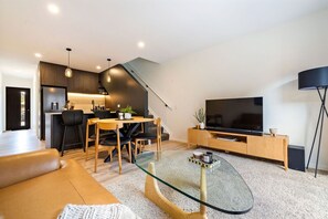 Una sala moderna con una mesa de centro de vidrio, un sofá, un comedor con sillas, una cocina al fondo y un televisor de pantalla plana sobre un soporte de madera.