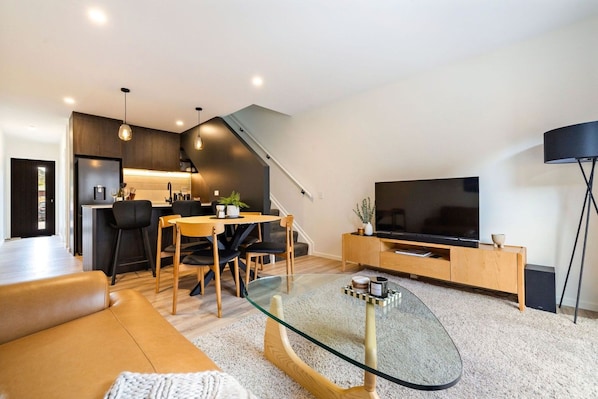 A modern living room with a glass coffee table, a sofa, a dining area with chairs, a kitchen in the background, and a flat-screen TV on a wooden stand. - Prime Location with mountain views (Queenstown)