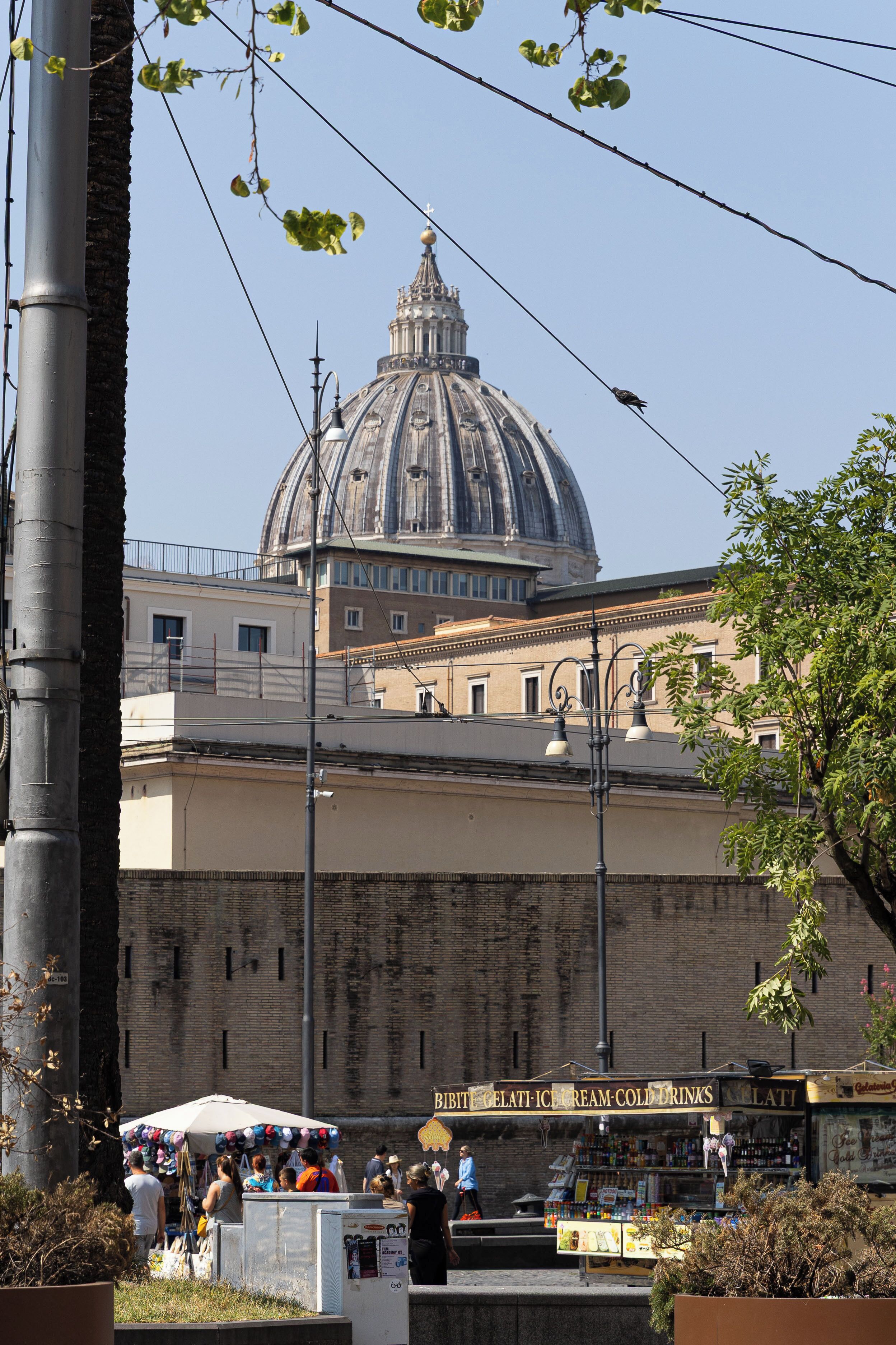 A street scene with a large dome building in the background, a market stall with a sign reading 'BITF GELATI-ICE CREAM-COLD DRINKS,' and a tree in the foreground.