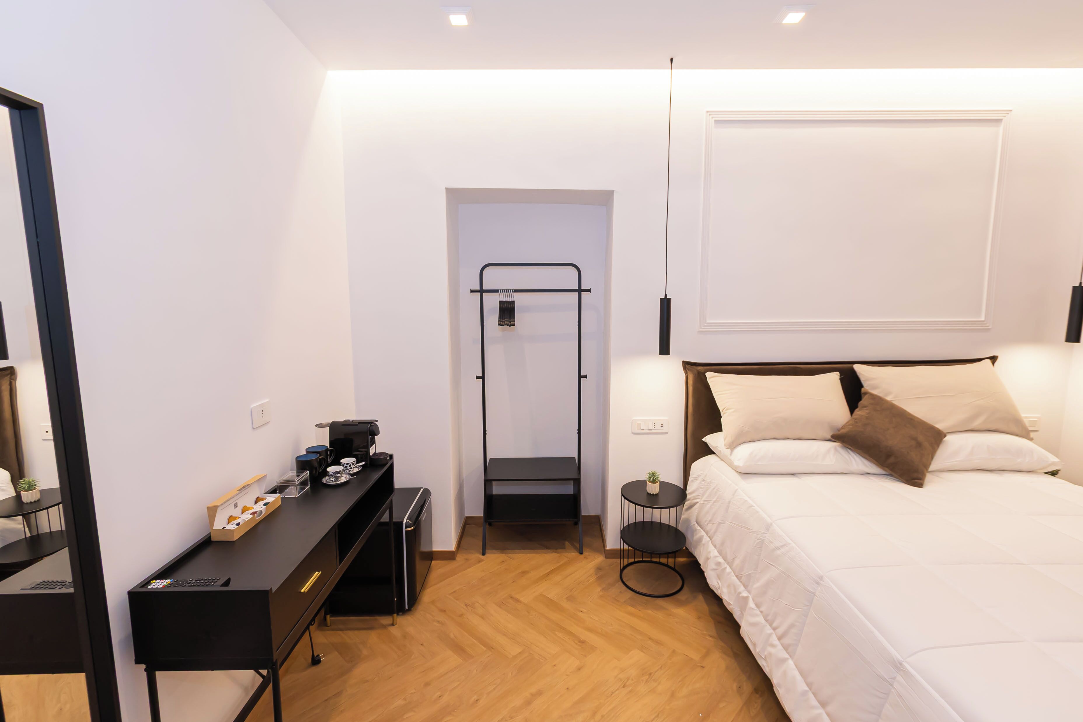 A hotel room with a bed, a desk, a chair, a small table, a mirror, and a wall-mounted shelf.