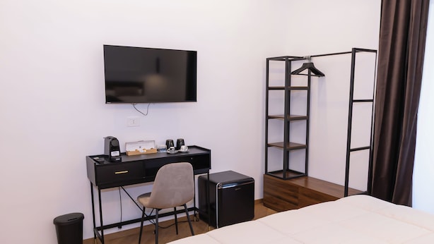 A hotel room with a bed, a desk with a chair, a television, a small black speaker, a trash bin, and a wardrobe with a mirror.