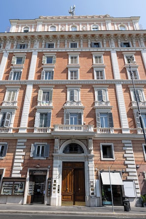 A multi-story building with a classic architectural style, featuring a central entrance with a wooden door, multiple windows with shutters, and a sign for a business on the ground floor. - Sweet Home 36-Vatican (Rome)