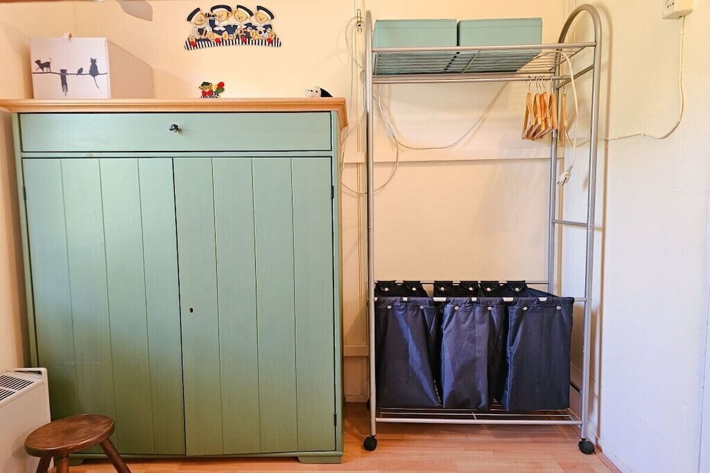 A room with a green wardrobe, a metal drying rack with clothes, and a small stool.
