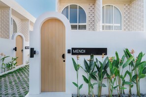 Un edificio moderno con puerta de madera, un cartel que dice 'MENDI' y plantas tropicales en primer plano.