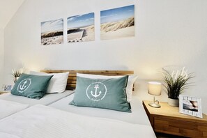 Ein Hotelzimmer mit einem Bett, zwei Kissen mit Anker-Motiven, einem Nachttisch mit Lampe, einem gerahmten Foto und Wandbildern mit einer Strandansicht.