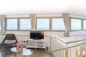 A room with a television on a white stand, a glass table with two candles, a wicker chair, and a view of the sea through large windows. - Exclusive tower apartment (Burgtiefe)