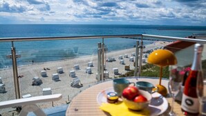 Un balcón con vista a una playa y al océano, una mesa con frutas y una botella, y una lámpara de sol.