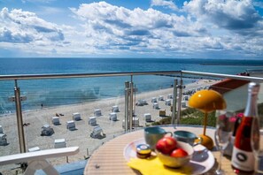A balcony with a view of a beach and ocean, a table with fruits and a bottle, and a sun lamp. - Exclusive tower apartment (Burgtiefe)