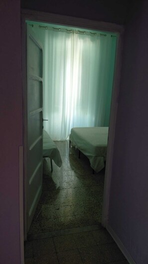 A narrow hallway with a bed and a wardrobe on one side and a window with curtains on the other.