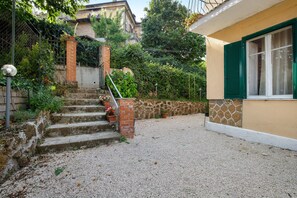 Un edifici residencial amb una escala de pedra que porta a una porta, envoltat de vegetació i un camí de grava.