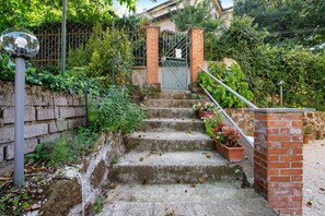 Un conjunt de graons de formigó que condueixen a una paret de maó amb una reixa metàl·lica, envoltat de vegetació i plantes en testos.