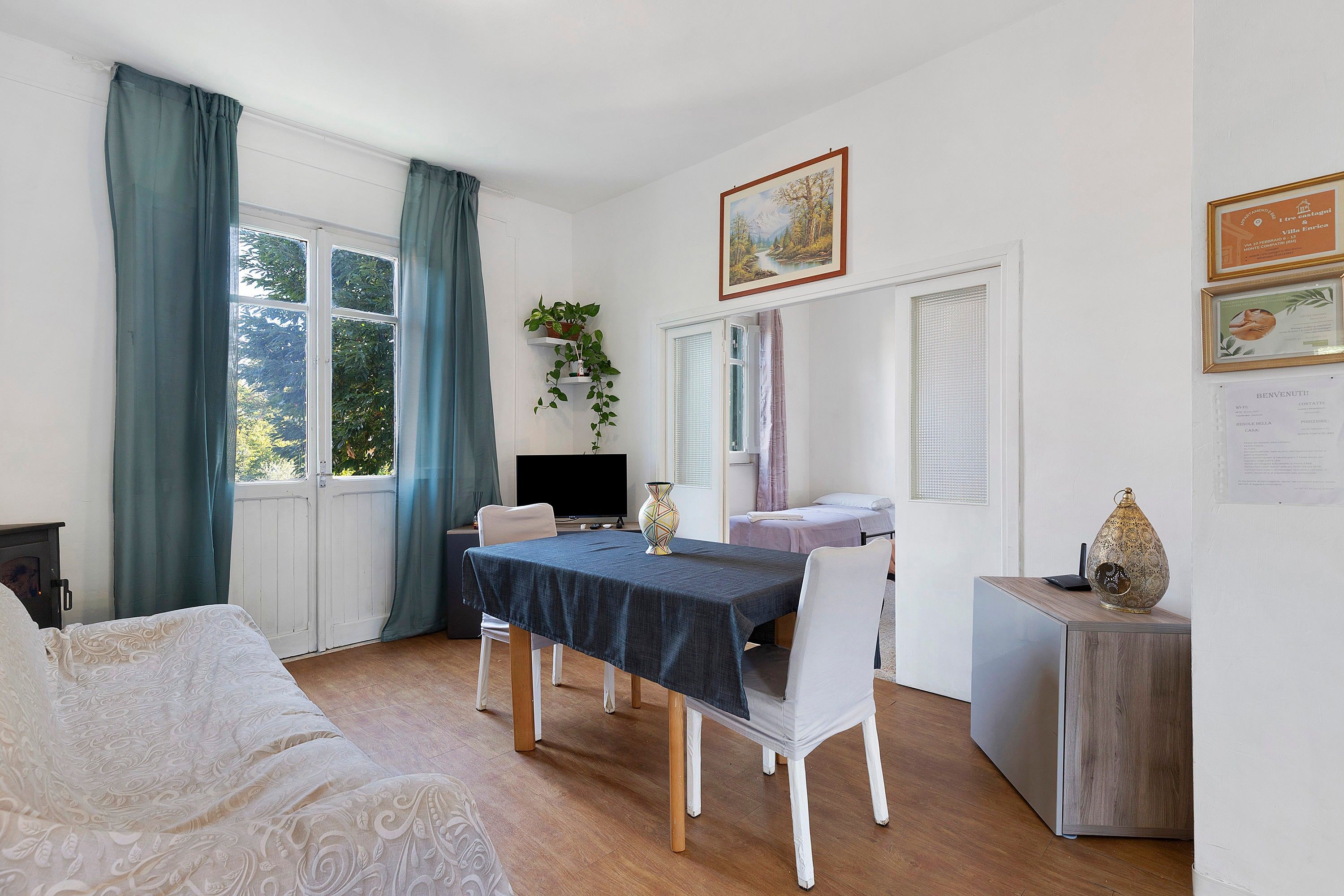 Una stanza con un letto, un tavolo con sedie, una TV e una finestra con tende.