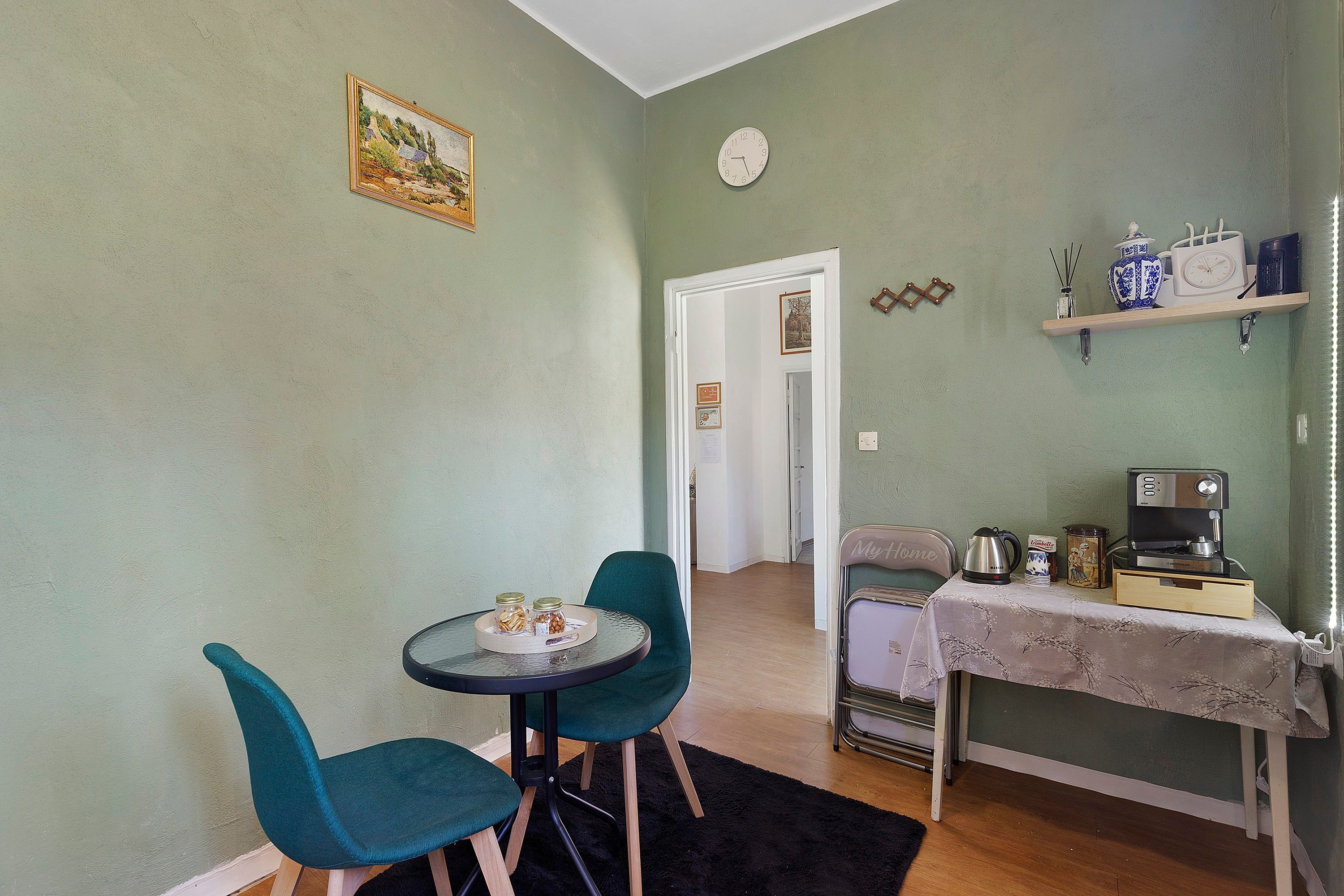 A small dining area with a table and two chairs, a kitchen area with a coffee machine, and a clock on the wall.