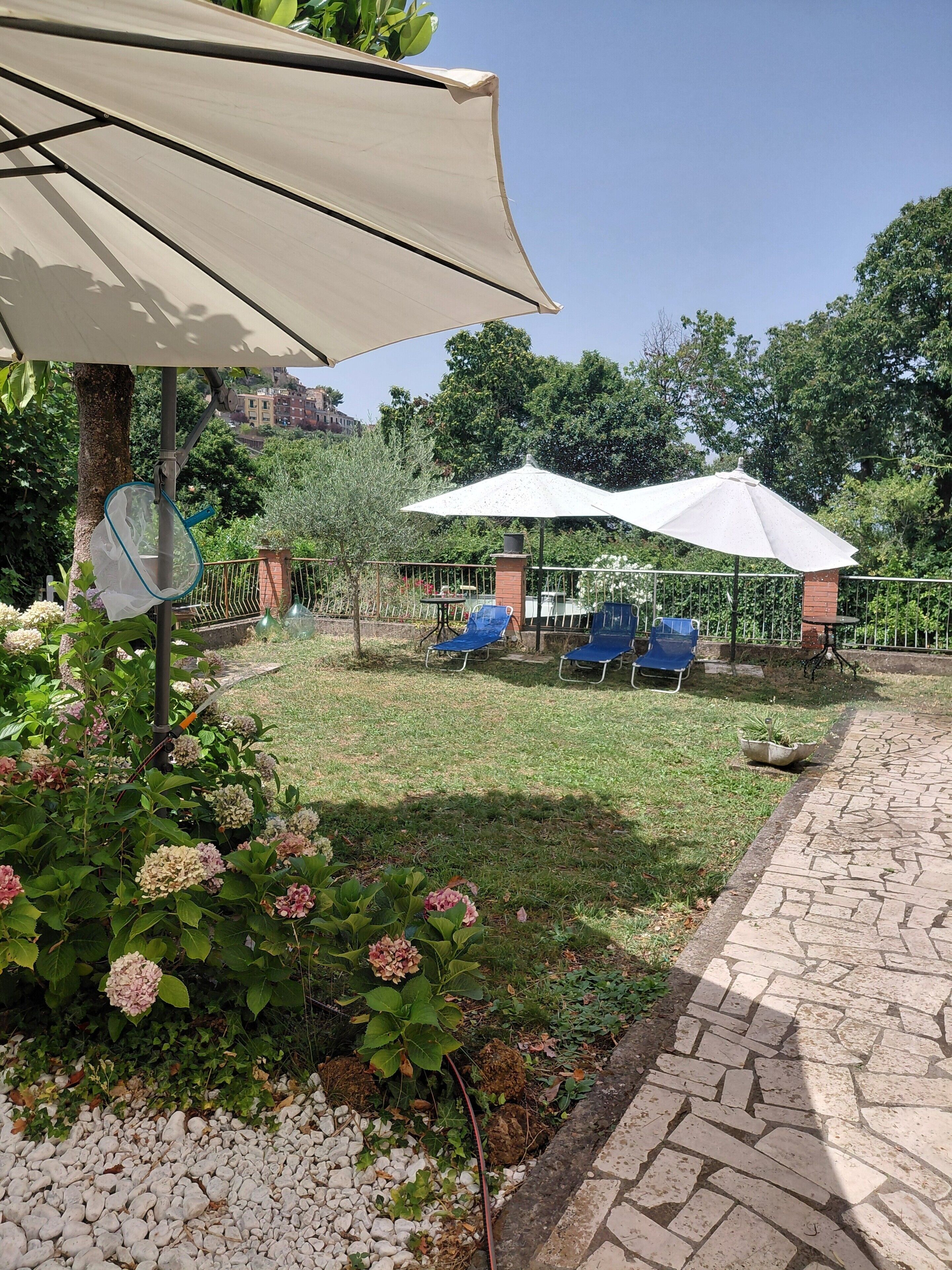 A garden area with a paved walkway, outdoor seating, and a pergola with a white umbrella.