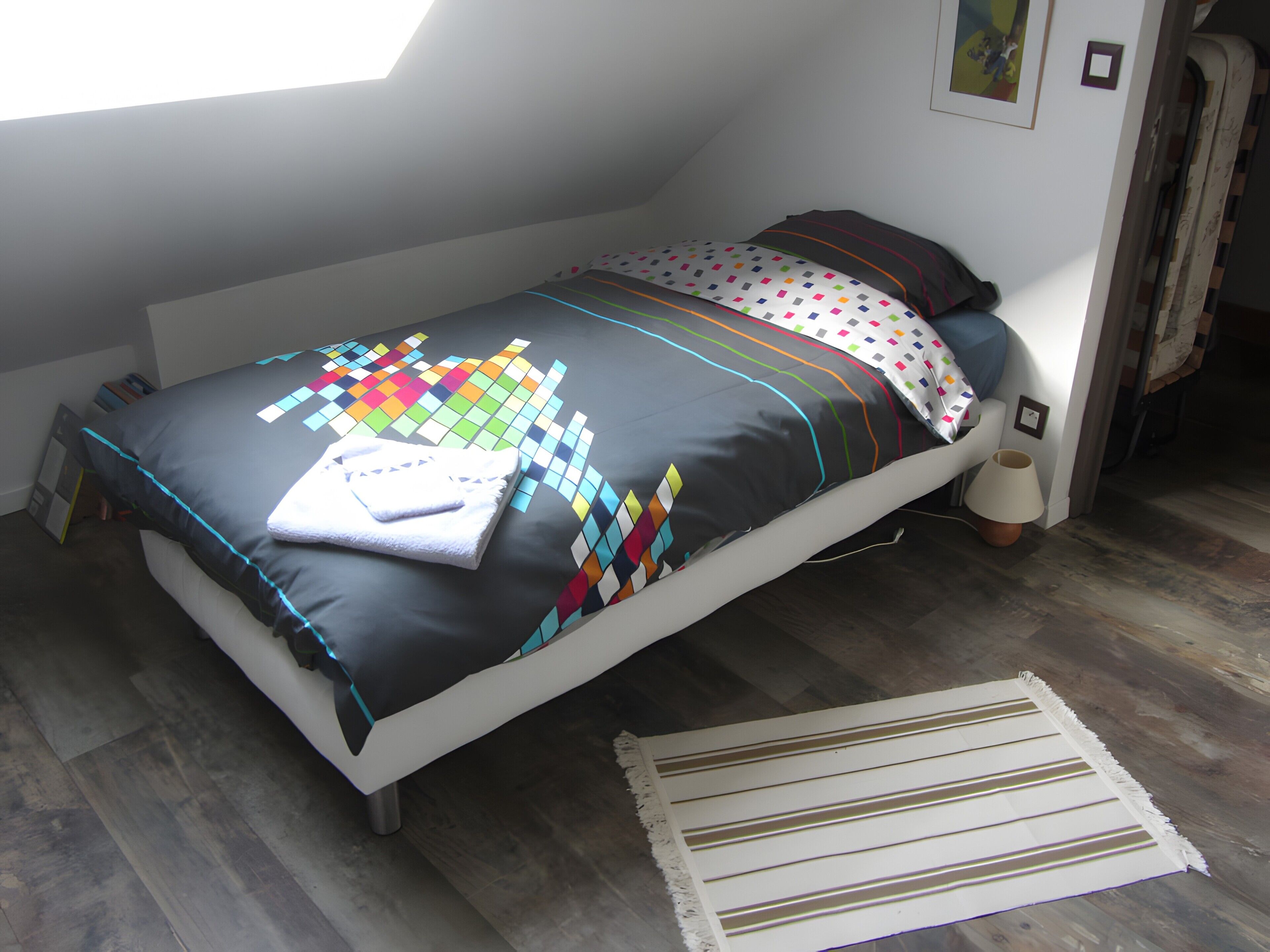Una cama individual con un edredón colorido y una almohada blanca, una mesita de noche con una lámpara, y una ventana con vistas al exterior.