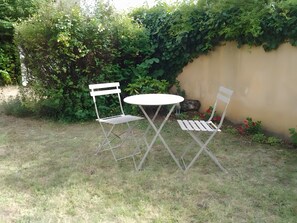 Ein kleiner Sitzbereich im Freien mit zwei Metallstühlen und einem runden Tisch, umgeben von Gras und Grünpflanzen.