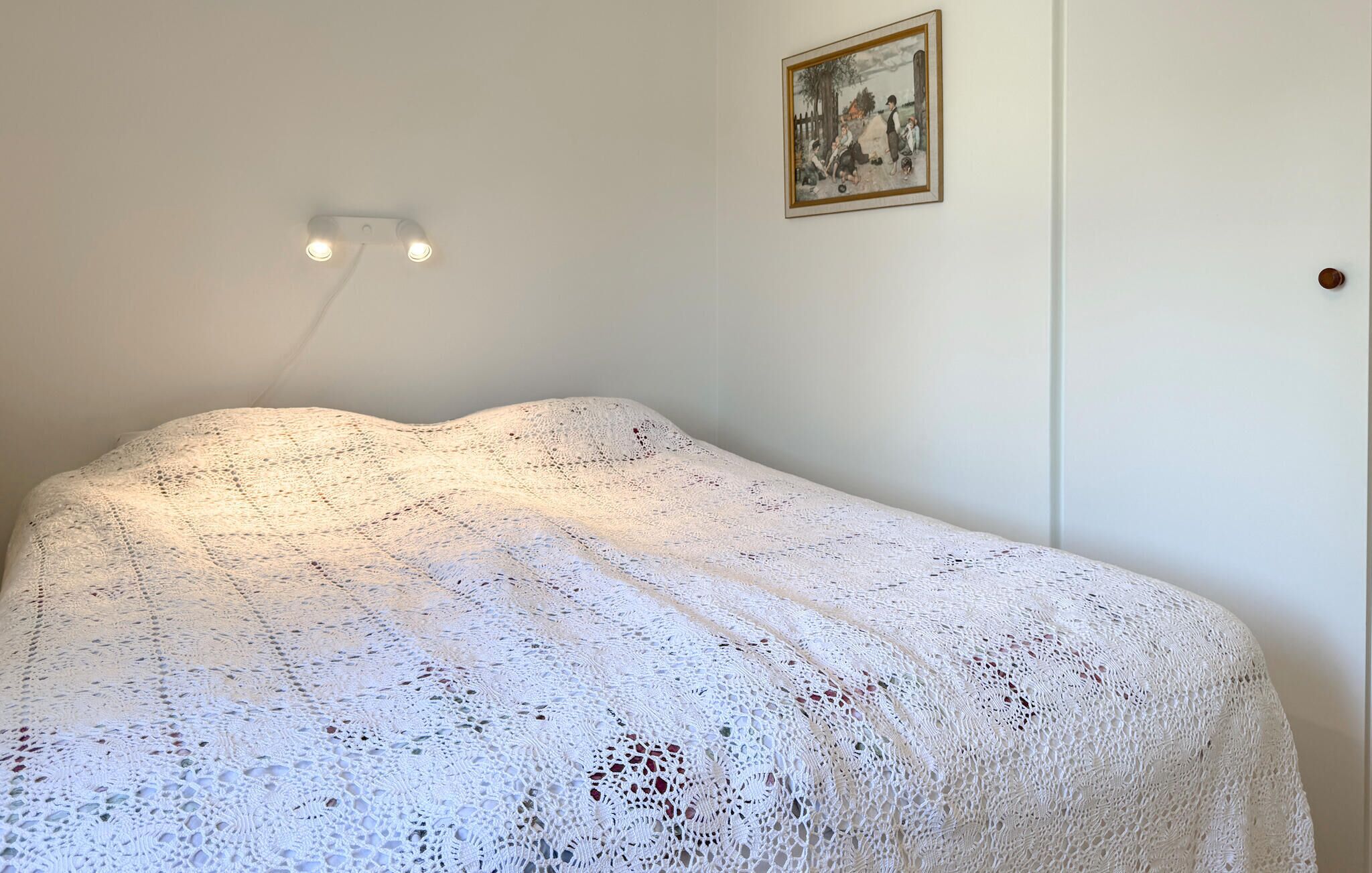 A room with a bed covered in a white knitted blanket, a wall-mounted light fixture, and a framed picture.